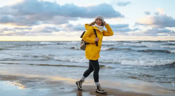 Uśmiechnięta kobieta w żółtej kurtce spaceruje po plaży. Wiatr rozwiewa jej włosy. To piękny jesienny dzień nad morzem.