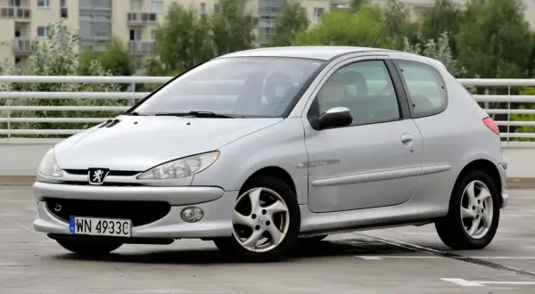 Srebrny Peugeot 206 z widocznymi przednimi żarówkami, zaparkowany na mokrym parkingu.