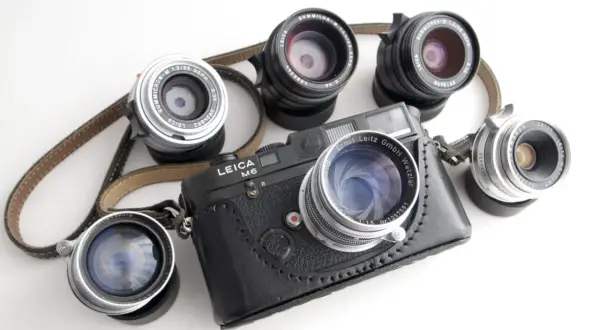 A Leica M6 camera with a leather case is surrounded by several best M mount lenses, showcasing a collection of classic photographic gear.