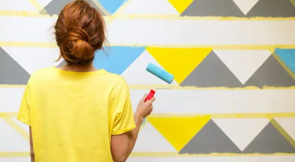Frau mit rotem Dutt streicht mit Roller geometrische Muster an Wand. Ideen für Wand streichen mit Muster abkleben.