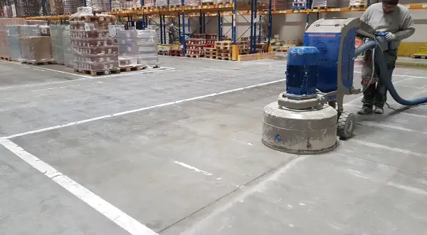 Pracownik wykonuje szlifowanie betonu w magazynie, używając maszyny z odkurzaczem.