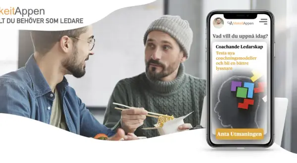 Två män äter lunch och pratar. En mobil visar appen MakeitAppen med en utmaning för coachande ledarskap, ett bra sätt att visa uppskattning på jobbet.