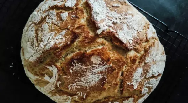 Złocisty, domowy bochenek chleba z chrupiącą skórką, posypany mąką, na czarnej kratce.