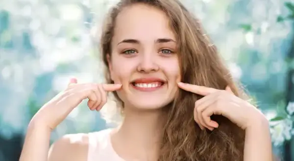 Uśmiechnięta dziewczyna z dołeczkami na policzkach, które podkreśla palcami. Wygląda jakby miała cheek piercing, ale to naturalne wgłębienia.