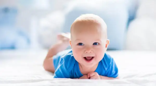 Uśmiechnięty 2 miesięczne dziecko w niebieskim body leży na brzuchu na białej pościeli.