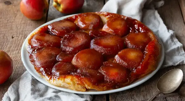 En gyllene tarte tatin med karamelliserade äpplen på en vit tallrik, omgiven av äpplen och en linneduk.
