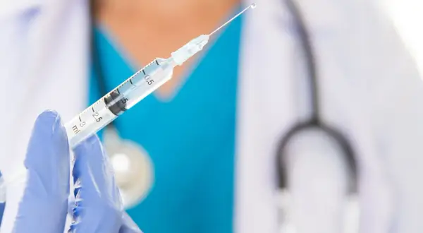 Lekarz w niebieskiej rękawiczce trzyma strzykawkę z kroplą szczepionki. Przygotowanie do szczepień.