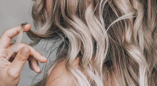 Dłoń dotyka falujących włosów w odcieniu sombre blond, podkreślając ich naturalne przejścia kolorystyczne.