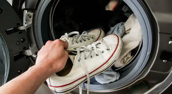 Mano che inserisce scarpe da ginnastica bianche in lavatrice per lavarle. La centrifuga le renderà come nuove!