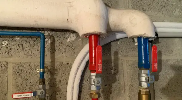 Tuyauterie gainée pour le calorifugeage, avec vannes rouges et bleues sur fond de mur en parpaings.