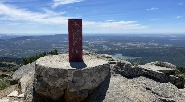 Pilar rojo con grafitis en la cima de una montaña, con un paisaje de valle y lago al fondo. La **cabeza lijar** del pilar parece desgastada por el tiempo.