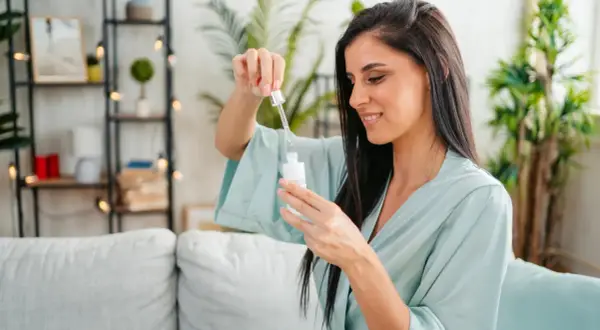 Frau mit dunklen Haaren in einem hellblauen Morgenmantel, die einen Tropfen eines Vitamin C Serums aus einer Pipette auf eine Flasche fallen lässt.