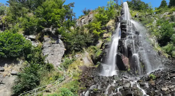 Der Trusetaler Wasserfall stürzt über schwarze Felsen in die Tiefe. Ein weißer Geländerweg führt Besucher am grünen Hang entlang.