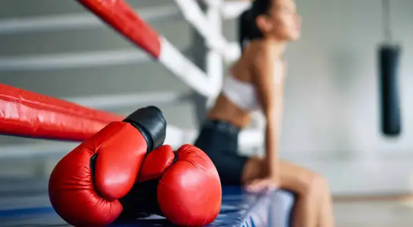 Red boxing gloves rest on the edge of a boxing ring. A woman sits in the background, perhaps contemplating how to do a body shot.