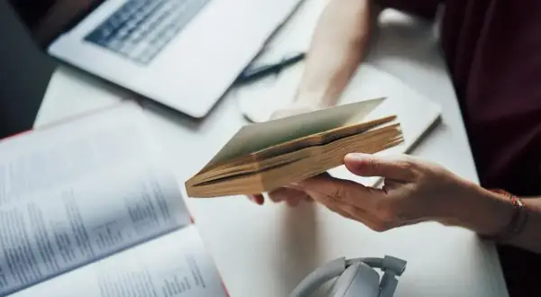 Ręce trzymają otwartą książkę, obok laptop i słuchawki. To dobry przykład, jak tworzyć bibliografię.