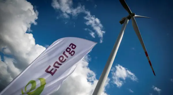 Flaga z logo Energa powiewa na tle błękitnego nieba z chmurami, obok wiatraka. Akcje Energa rosną.