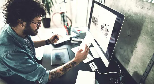 Ein Mann mit lockigen Haaren und Tattoos arbeitet an seinem Computer. Er ist im Bereich IU Kommunikationsdesign tätig und entwirft visuelle Konzepte.