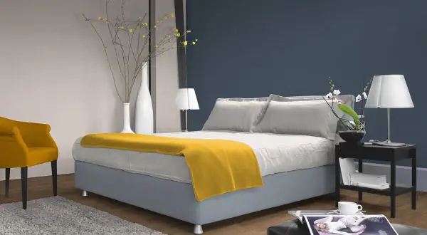 Ein Schlafzimmer mit einem Bett, das mit einer gelben Decke und Kissen in verschiedenen **blau nuancen** dekoriert ist. Ein gelber Sessel und ein Teppich ergänzen den Raum.