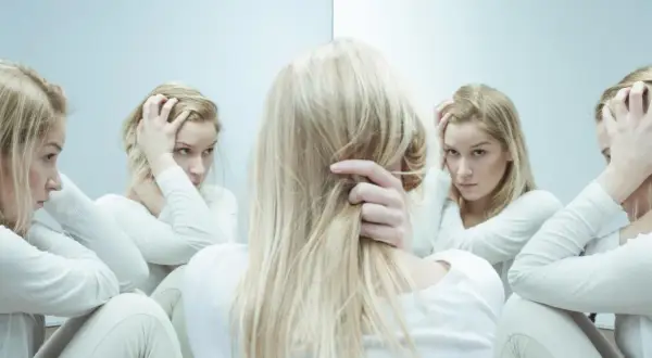 Kobieta w białej bluzie z długimi blond włosami, zmagająca się z kwetaplex, widzi swoje odbicia w lustrze, każde z innym wyrazem twarzy.