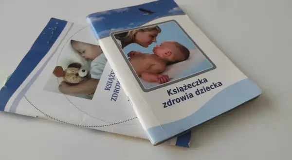 Dwie książeczki zdrowia dziecka leżą na białym tle. Jedna z nich przedstawia matkę całującą niemowlę, druga śpiące dziecko z misiem.