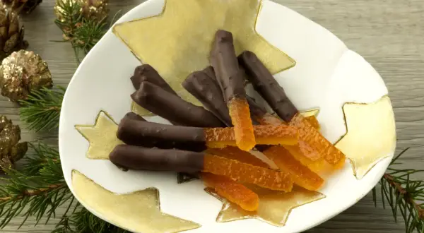 Bâtonnets d'orange confite chocolatés sur une assiette étoilée, entourés de pommes de pin dorées et de branches de sapin.