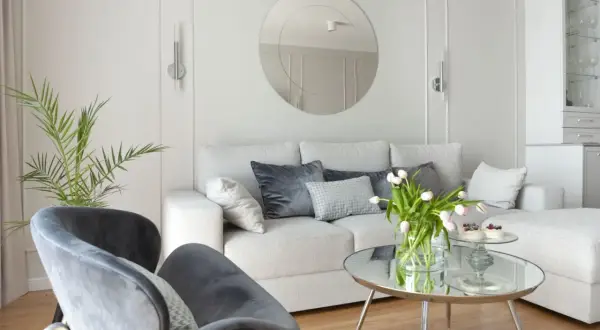 Przytulny salon z jasnoszarymi, **białymi ścianami**, wygodną kanapą, welurowymi fotelami i okrągłym lustrem.