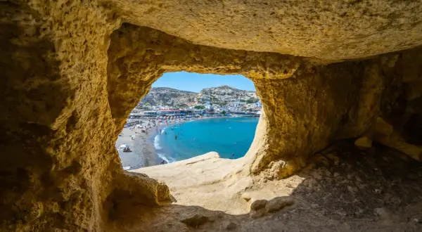 Blick durch eine Höhle auf einen belebten Strand und das türkisfarbene Meer. Ein perfekter Ausschnitt der Kreta Highlights.