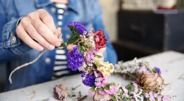 Hände binden bunte Trockenblumen zu einem Kranz. Ein schönes Projekt, um Trockenblumen selbst zu machen.