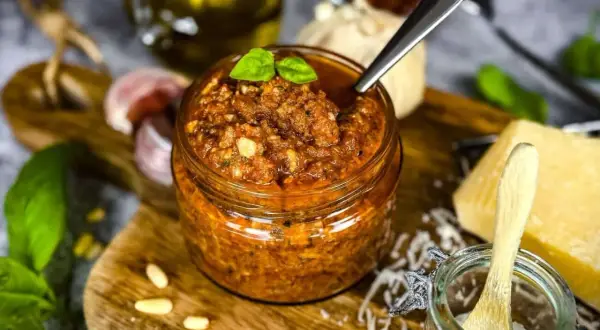 Ein Glas mit leuchtend rotem Pesto, garniert mit Basilikum. Daneben liegen Pinienkerne, Knoblauch und ein Stück Parmesan.