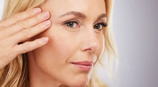 Mujer con piel madura, tocando suavemente su rostro, mostrando líneas de expresión y una mirada serena.