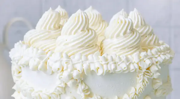 Eine üppige Buttercreme Torte mit kunstvollen Tupfen und Rüschen aus weißer Buttercreme, die auf einem weißen Hintergrund steht.