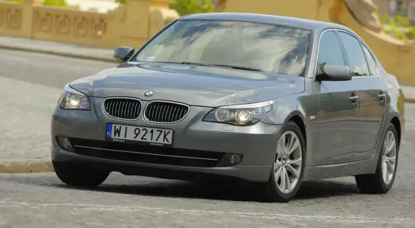 Szary sedan BMW E60 z włączonymi światłami. Widać charakterystyczne żarówki BMW E60, które oświetlają drogę.