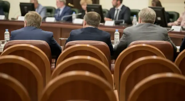 Mężczyźni w garniturach siedzą przy stole, czekając na **absolutorium**. W tle widać monitory i innych uczestników spotkania.
