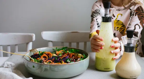 En färgstark sallad med morötter och rödkål i en skål, bredvid två flaskor med dressing till sallad. Ett barn håller i den ena flaskan.