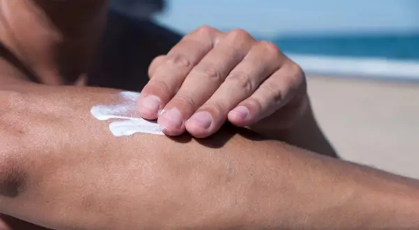 Dłoń rozsmarowuje krem z filtrem, zawierający m.in. diethylamino hydroxybenzoyl hexyl benzoate, na ramieniu na plaży.