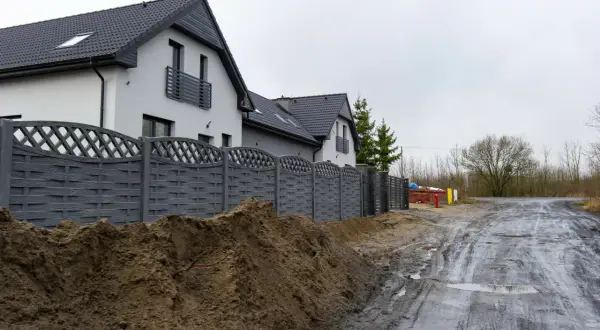 Nowy, szary płot dzieli posesje, sugerując konflikt z sąsiadem o ogrodzenie. Błotnista droga i sterta ziemi podkreślają napięcie.