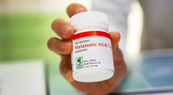 En hand håller en burk med 100 melatonin tabletter. Frågan "hur mycket melatonin kan man ta" kan besvaras genom att läsa etiketten.