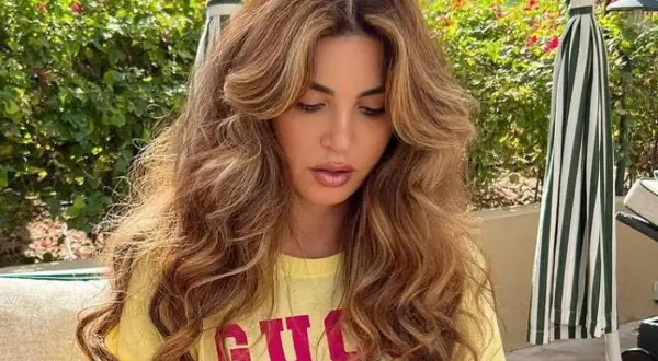 Mujer con cabello largo y ondulado, con un corte mariposa que enmarca su rostro. Viste una camiseta amarilla con letras rosas.
