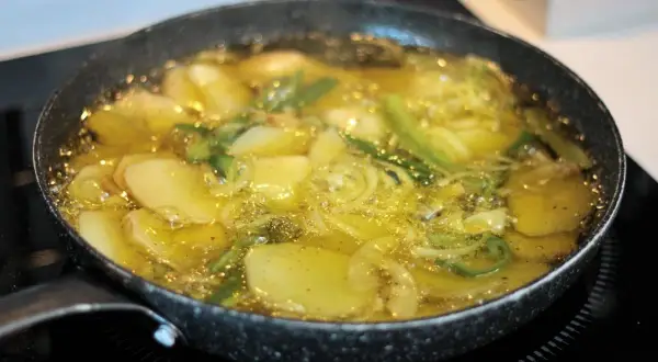 Patatas panaderas en sartén, dorándose en aceite caliente con cebolla y hierbas.