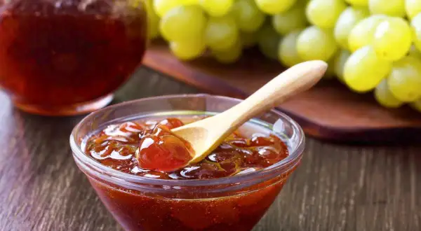 Un cuenco de cristal con mermelada de uvas casera, una cuchara de madera y un racimo de uvas verdes al fondo.