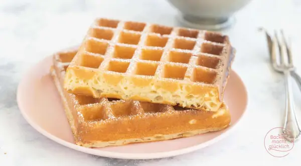 Zwei goldbraune belgische Waffeln, mit Puderzucker bestäubt, auf einem rosa Teller.