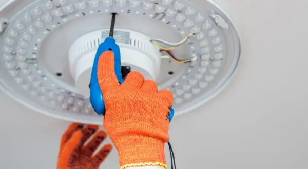 Ręka w pomarańczowej rękawicy przykręca śrubokrętem lampę sufitową LED. Dowiedz się, jak zrobić oświetlenie.