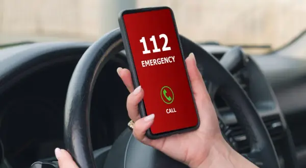 Mano sujetando un teléfono con pantalla roja que muestra "112 EMERGENCY" y un botón de llamada verde. El volante del coche está visible.