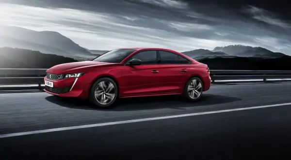 Voiture rouge Peugeot 508, un modèle à éviter, roulant sur une route avec des montagnes en arrière-plan sous un ciel dramatique.
