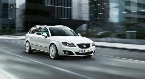 Biały Seat Exeo 2.0 TDI kombi pędzi ulicą miasta, mijając nowoczesne budynki.