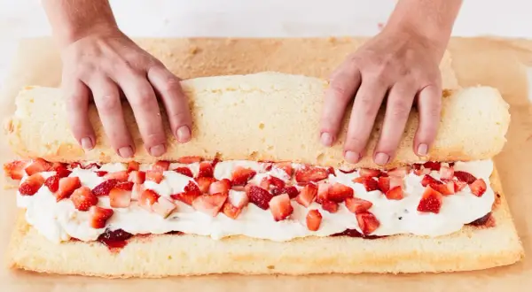 Hände rollen einen Biskuitteig mit Sahne und Erdbeeren für ein Erdbeerrolle Rezept auf.