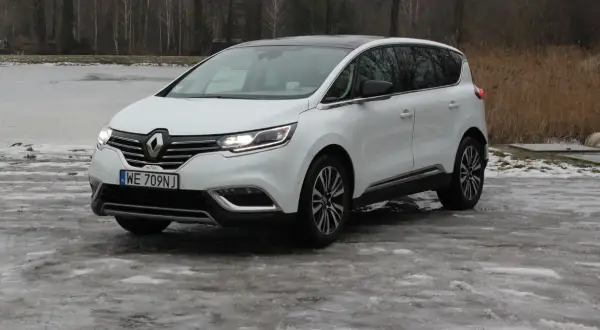 Biały Renault Initiale na oblodzonym podjeździe, z widokiem na zamarznięte jezioro i drzewa.