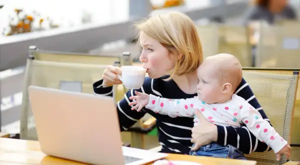 Kobieta pije kawę, trzymając dziecko. Obok leży laptop. Karmienie piersią i chwila dla siebie.