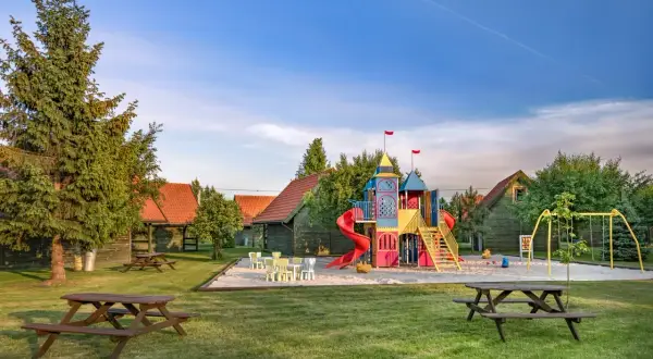 Kolorowy plac zabaw w kształcie zamku, huśtawki i stoły piknikowe na trawniku. W tle drewniane domki, idealne na łebska chata.