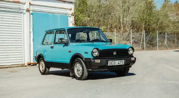 En ljusblå Lancia A112 Junior står parkerad framför en blå port och en vit containerbyggnad.